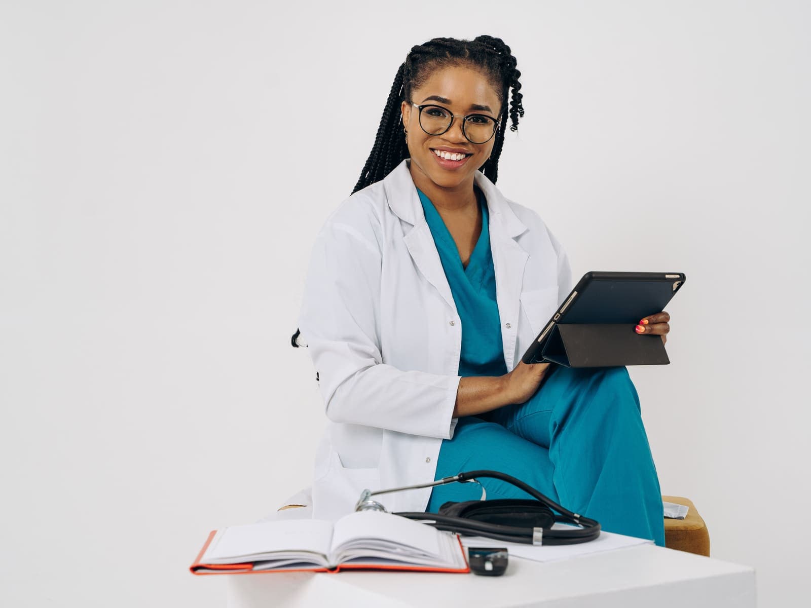 A doctor in a white coat and scrubs sitting with a tablet, a stethoscope and notes beside her.
